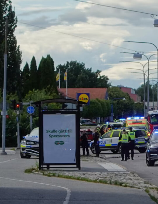 Polisinsats med blåljus och flera polisbilar och en ambulans vid en trafikolycka nära en skylt med texten "Skulle gått till Specsavers" och en Lidl-butik i bakgrunden.