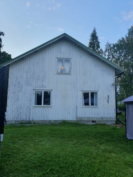 Vit träfasad på en byggnad med två fönsterbågar, varav en nyrenoverad på övervåningen. Gräsmatta och träd runt huset.