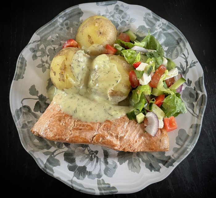 Stekt lax serverad med kokt potatis, grönsallad med tomater, gurka, och rödlök, toppad med smögensås på en blommönstrad tallrik.