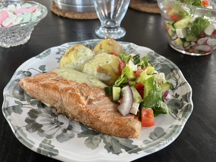 Tallrik med stekt lax, kokt potatis med sås och en färgglad sallad. I bakgrunden en skål med sallad och en skål med godis.