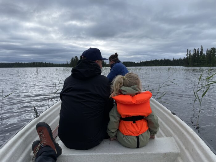 Personer sitter i en båt på en sjö en molnig dag, varav ett barn med flytväst tittar på vattnet.