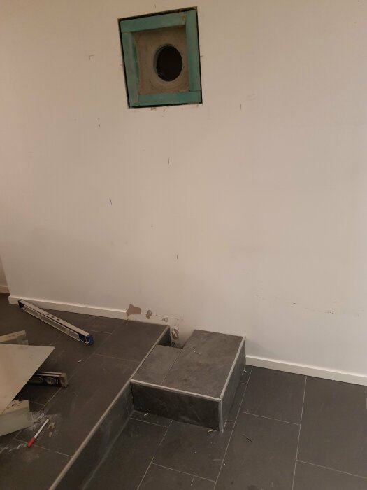 Installationsplats för kamin med ett hål i väggen för rökrör, nivåskillnad i golvet med trappa, verktyg och byggmaterial på mörkt klinkergolv.