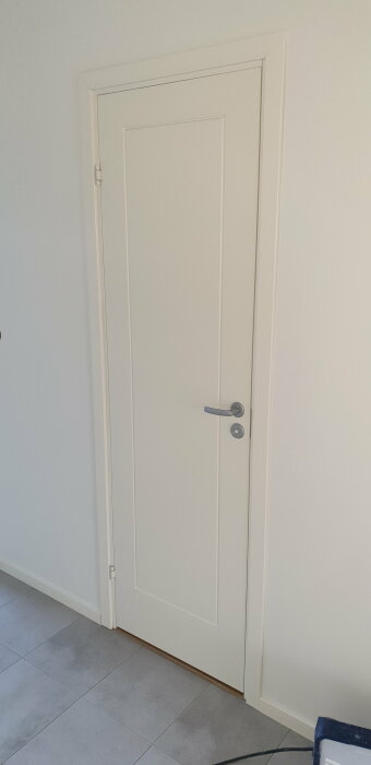 Vit dörr med silverfärgat handtag i vitmålad vägg, bredvid kaklat golv i en hall, som del av en totalrenovering av lägenhet.