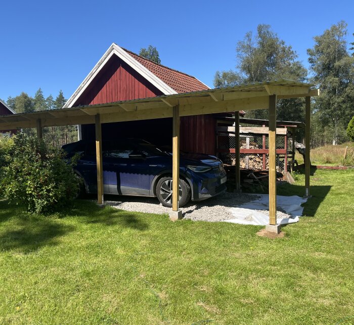 En carport under uppbyggnad, med stolpar och tak av trä bredvid rött hus, täckt med vit markduk och grus. En mörkblå bil står parkerad under taket.