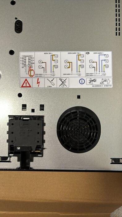 Närbild på baksidan av en häll med anslutningsinstruktioner och en liten svart fläkt. Instruktionsetikett visar elkonfigurationer och varningar.