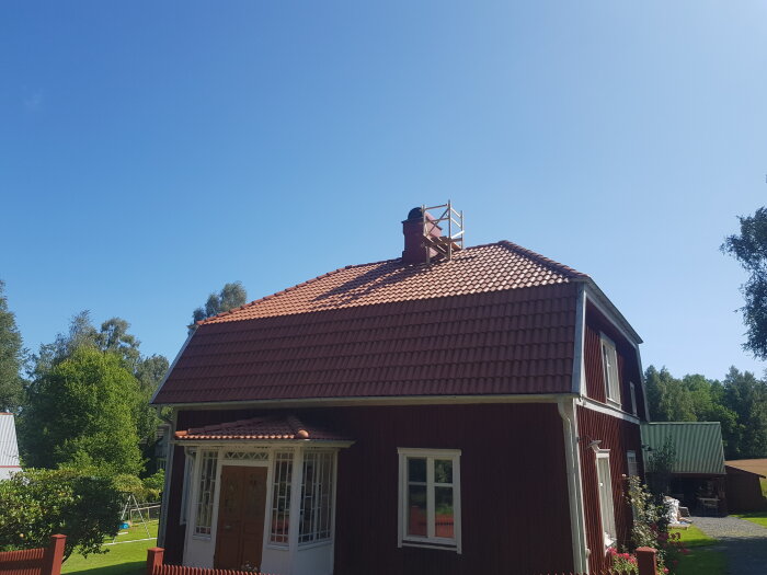 Rött hus med tegeltak och en trälåda byggd kring skorstenen, tydligen inspirerad av Alfons Åbergs helikopter. Strålande sol och grönskande omgivning.