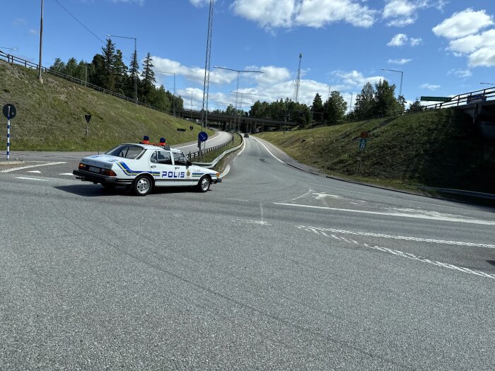 En gammal svensk polisbil står vid en korsning under en blå himmel med vita moln och omgivande grönska.