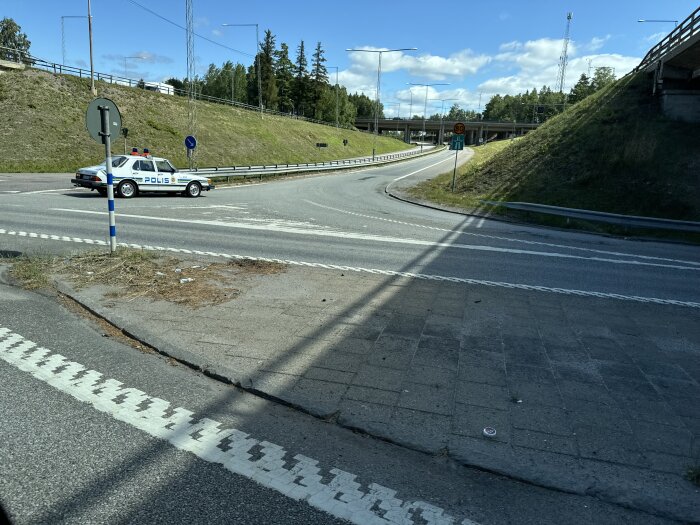 Polisbil blockerar en väg till en avfart, omgiven av asfalt och betongblock, med skog i bakgrunden under en solig dag.