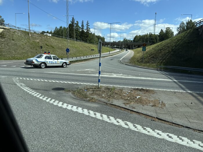 En polisbil står i en T-korsning på en öppen väg med trafikskyltar och en bro i bakgrunden under en klar himmel.