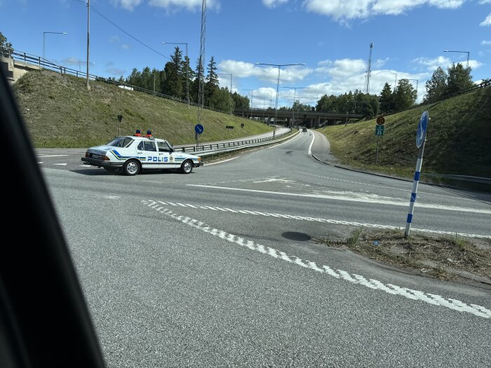 En polisbil blockerar vägen vid en motorvägskorsning under en klarblå himmel och grönskande omgivning.