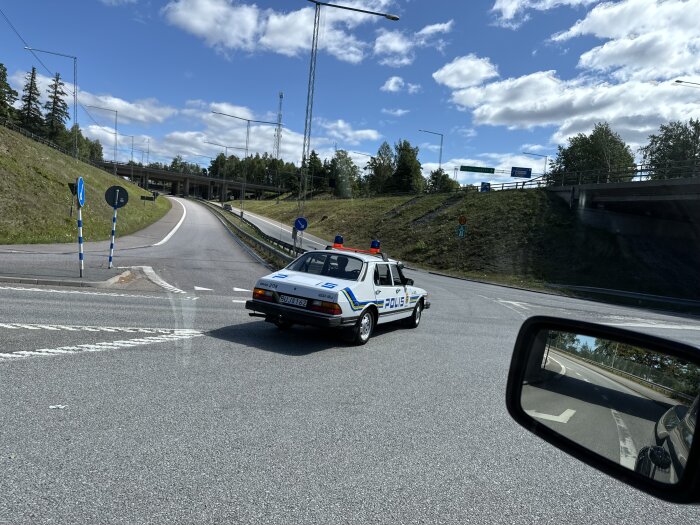 Polisbil vid korsning nära motorväg, som fotats från en bil. Klarblå himmel och grönska i bakgrunden.