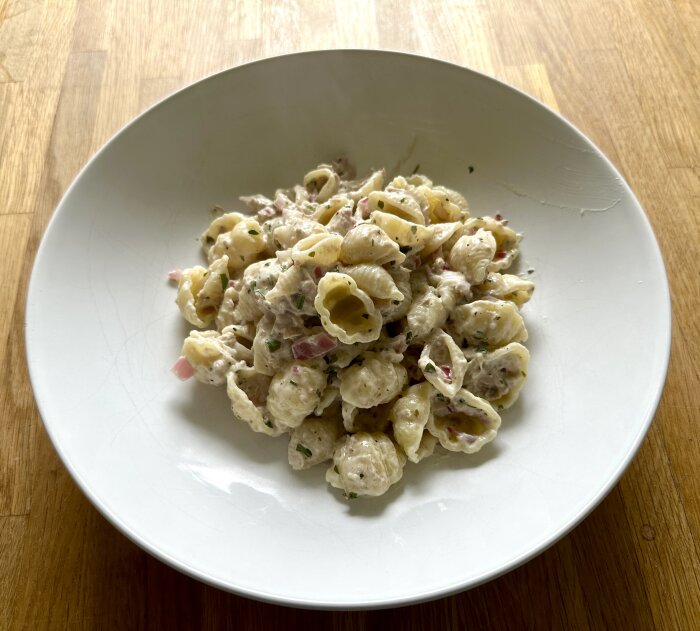 Vit tallrik med tonfiskpasta på ett träbord. Pastan är uppblandad med en krämig sås och hackad lök.