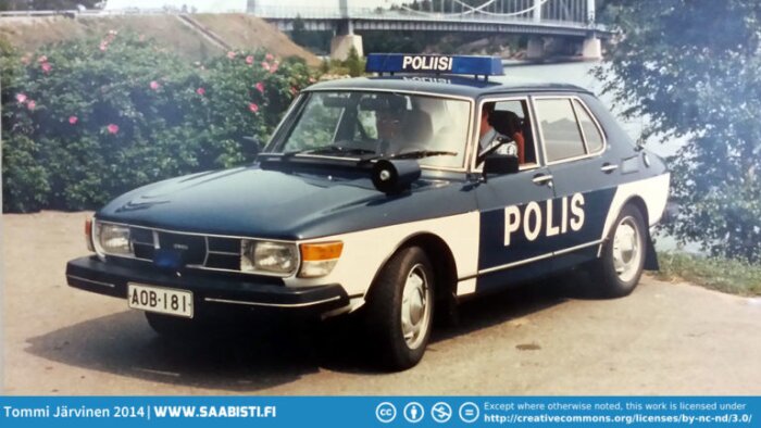 En Saab 99 med finsk polisbilsmärkning står parkerad vid vägkant, med bro och blommande buskar i bakgrunden.