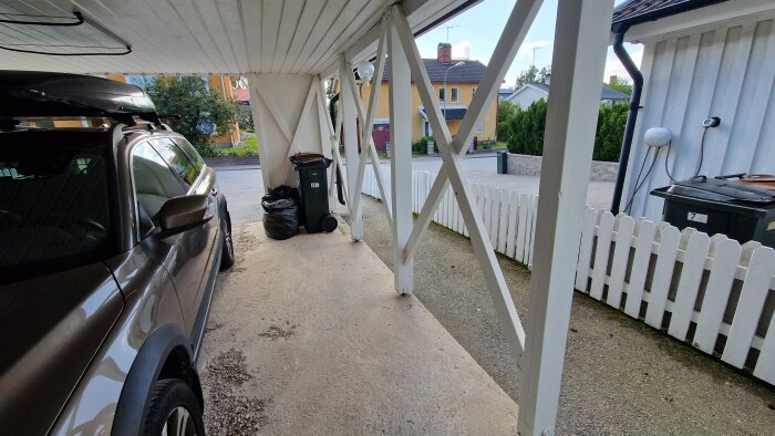 Bild av en carport med "kryss" mellan stolparna, en parkerad bil, soptunnor och vita trä-staket på sidorna.