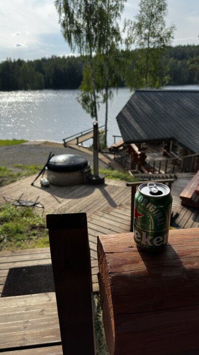 En utsikt över en sjö med skog i bakgrunden, en altan med en Heineken-burk på räcket, och en badtunna på trädäcket.