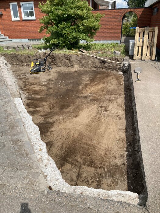 Uppgrävd mark bredvid ett hus med en gräsmatta och en träpall i bakgrunden. En komprimeringsmaskin och spade syns på platsen.