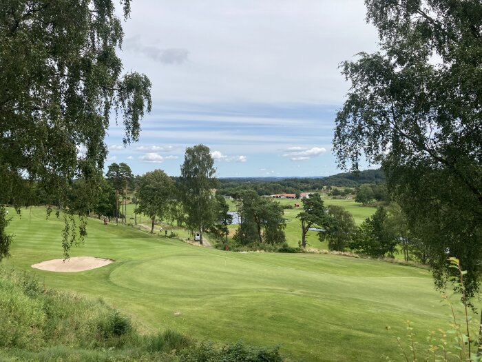 Vy över en grön golfbana med sandbunker i förgrunden, omgiven av träd. Blå himmel med några moln i bakgrunden och en röd byggnad i fjärran.