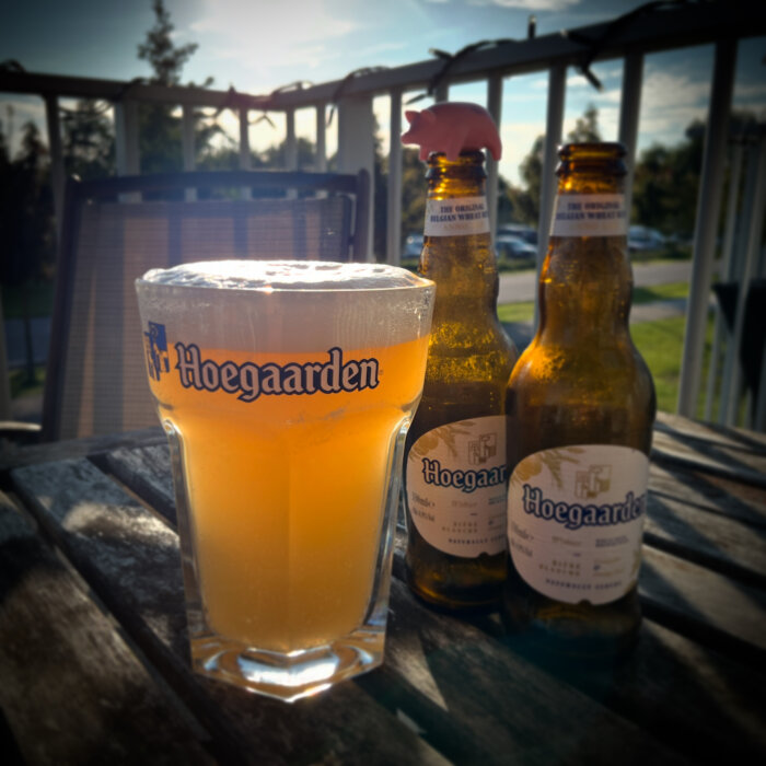 Ett glas Hoegaarden öl och två Hoegaarden ölflaskor på ett bord utomhus i solsken. En plastgris syns på en av flaskorna.