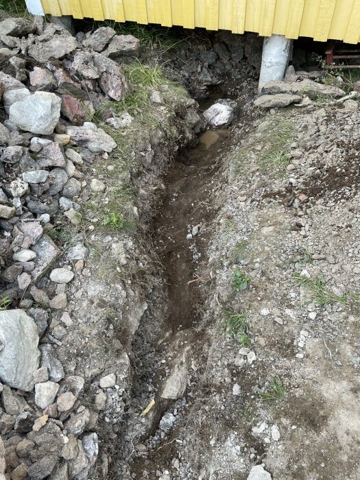 Dike grävt intill ett hus på plintgrund som står på berg, omgiven av sprängsten och jord, med en liten vattenpöl under huset nära berget och grunden.