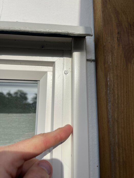 En person pekar på en vit plåtlist monterad runt en byggnadsfönsterkarm med vita väggar och träpanel i närheten.