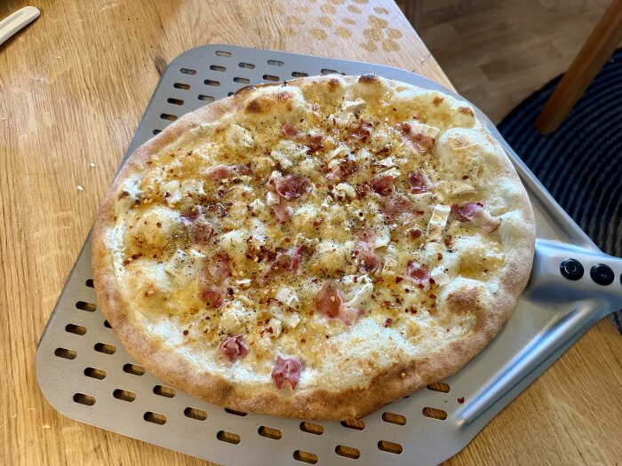 Pizza med bacon, chèvreost och honung på tunn, färdig deg, liggande på en perforerad pizzaplåt på ett träbord.