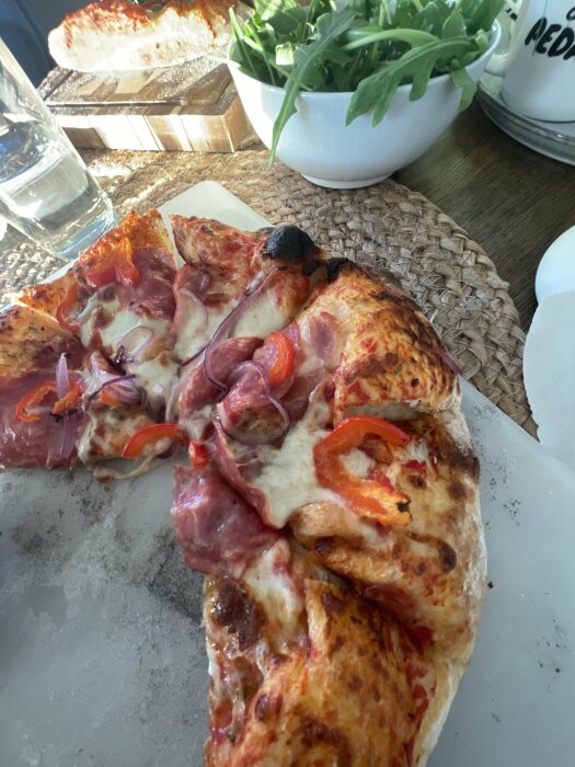 Hemlagad pizza med salami, paprika och lök på ett bord på en flätad bordstablett, med ett glas vatten och en skål med ruccola i bakgrunden.