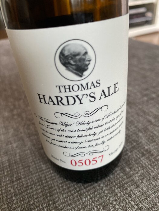 Flaska med etiketten "Thomas Hardy's Ale" och text om ölets kvalitet. Flasknummer 05057. Flaskans etikett är vit med svart text och en grå bild av en mans profil.