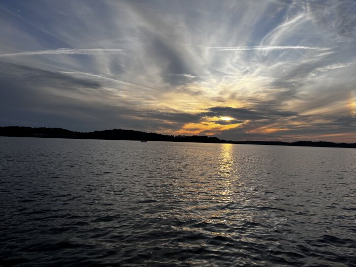 Solnedgång över en sjö med reflekterande vattenytor och silhuett av skogsklädda kullar i bakgrunden, samt moln som sprider sig över himlen.