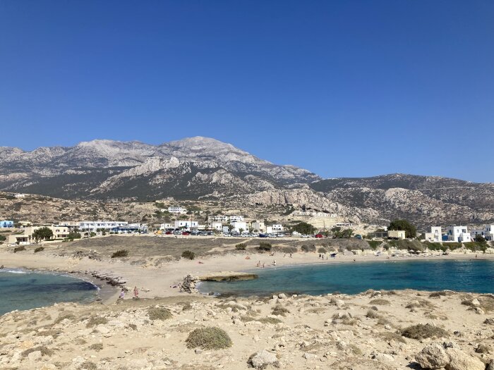 Strand med klippor och klart blått vatten i förgrunden, små vita hus och bilar i en by på kusten och berg i bakgrunden under en klarblå himmel.