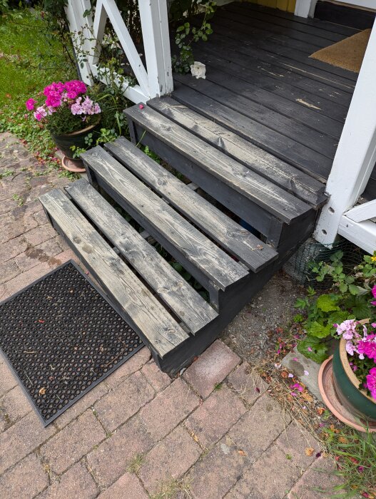 Slitna trätrappsteg och veranda med mörkt golv, omgärdad av blommor och grönska. En svart dörrmatta placerad vid trappans fot.