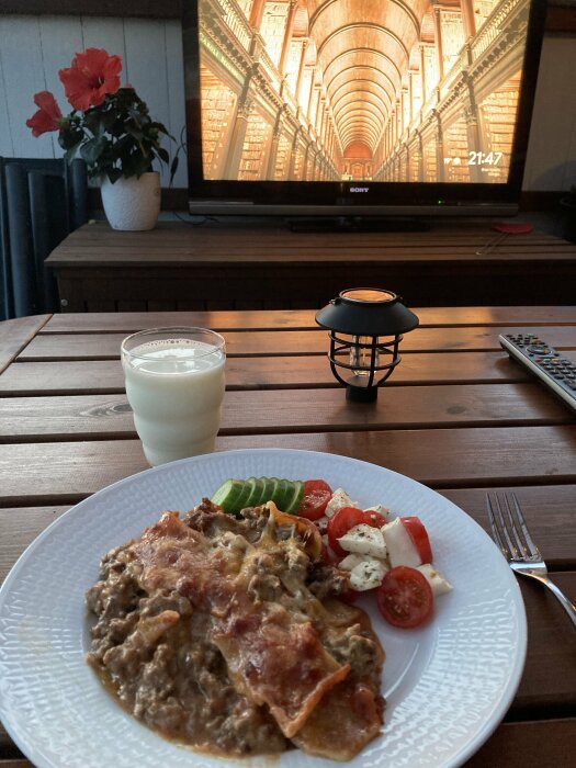 En tallrik med lasagne och grönsakstillbehör, ett glas mjölk och en TV i bakgrunden, med en bild av en lång sal fylld med böcker.