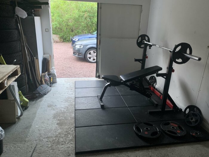 Garagegym med en bänkpress, viktskivor på golvet och en stång med vikter. Garageporten är öppen och en bil syns utanför på en grusuppfart.