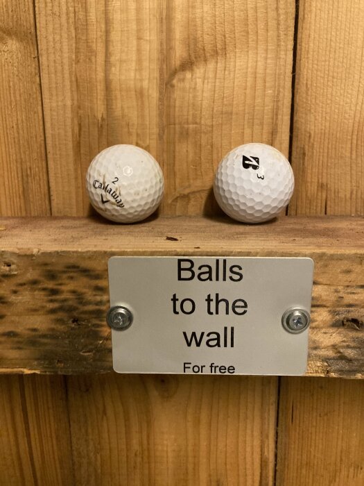 Två golfbollar sitter fast på en trävägg ovanför en skylt med texten "Balls to the wall For free".