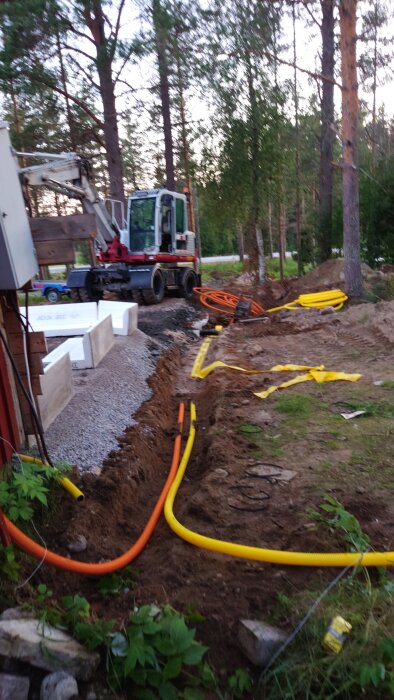 Grävmaskin vid ett hus, grävt dike med nerlagda orange och gula kablar för el och framtida fiber, omgiven av träd i en skogsmiljö.