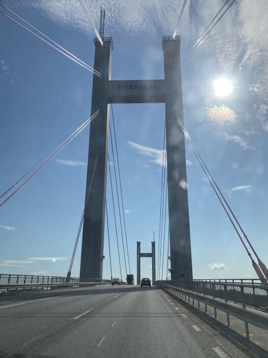 Bild på Tjörnbron med klar himmel, synlig trafik på bron med ett par bilar, vajrar och pelare som sträcker sig uppåt, sol synlig på himlen.