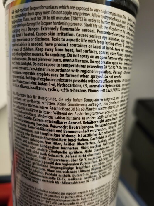 Närbild av en sprayburk etikett med instruktioner för användning av värmetålig lackfärg, som kräver härdning i 180 grader i 30-60 minuter. Innehåller säkerhetsvarningar.