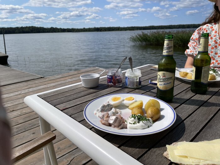 Sommarbord med mat bestående av kokta ägg, potatis och inlagd sill samt alkoholfri ölflaska, placerat vid en sjö med brygga i bakgrunden.