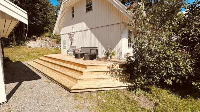 Nybyggd terrass av trallvirke med stegomslutning, fäst vid en vitmålad villa. Två svarta stolar och ett bord står på terrassen omgivna av grönska.