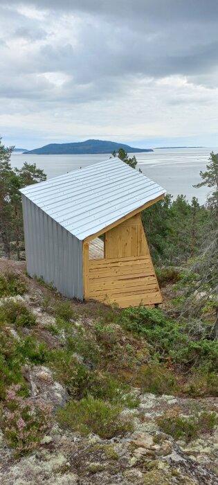 Liten stuga under uppbyggnad med plåttak och träväggar, belägen på ett berg med utsikt över en sjö och skog i bakgrunden.