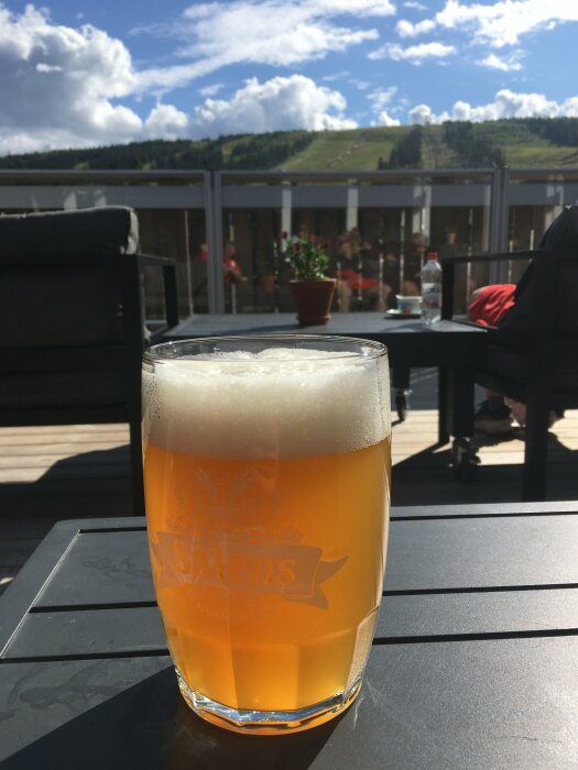 Ett glas öl med skum på toppen står på ett bord utomhus. I bakgrunden syns en stol och en blomkruka, samt en grönskande kulle under en blå himmel.