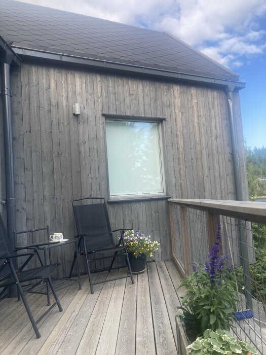 Fasad av träpanel på ett hus med en liten uteplats, två svarta stolar, ett bord med en kaffekopp, samt blommor i krukor.