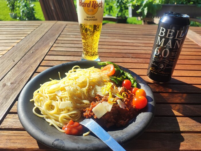 En tallrik spaghetti bolognese med ost, körsbärstomater och chili, med en Bellman-öl och ett glas öl på ett träbord i trädgården.