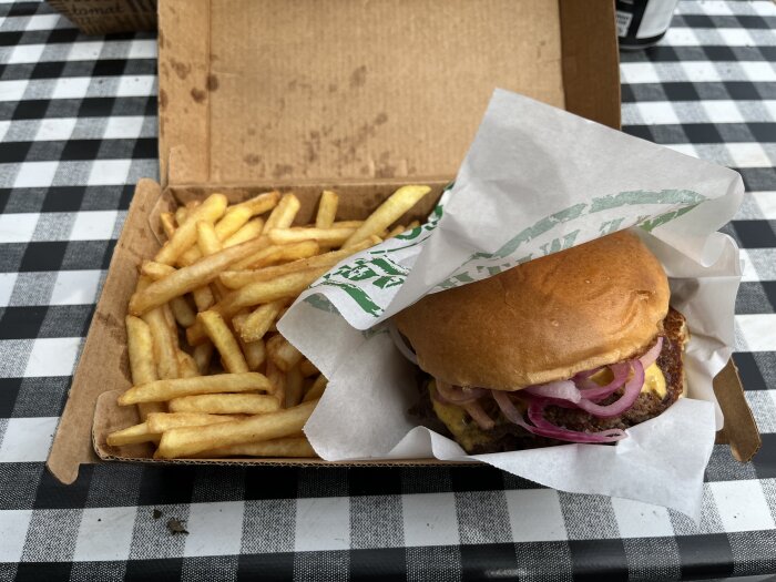 Hamburgare med ost och lök samt pommes frites, serverade i en kartong för avhämtning på ett svartvitt rutigt bord.