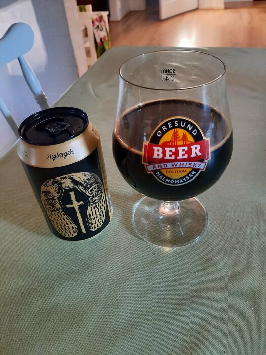 Bild på en ölburk från Stigbergets och ett glas fyllt med mörk öl, placerade på ett bord. Glaset har en etikett från Öresund Beer and Whisky Festival.