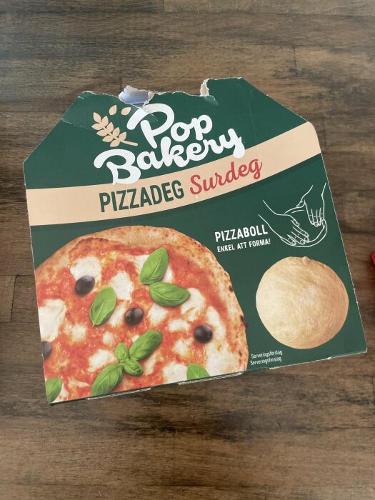 Förpackning av Pop Bakery pizzadeg surdeg på trägolv. Bild innehåller en pizza med mozzarella, basilika och oliver, samt instruktioner för pizzaboll.