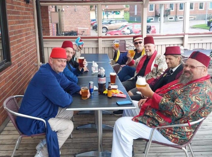 En grupp män i färgglada kläder och fez-hattar sitter runt ett bord utomhus, håller upp och skålar med glas fyllda med öl och ser glada ut.