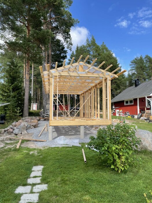 Stommen till en byggnad med väggblock och takstolar under en klarblå himmel. Byggnaden står på en upphöjd grund med grönska och stenar i bakgrunden.