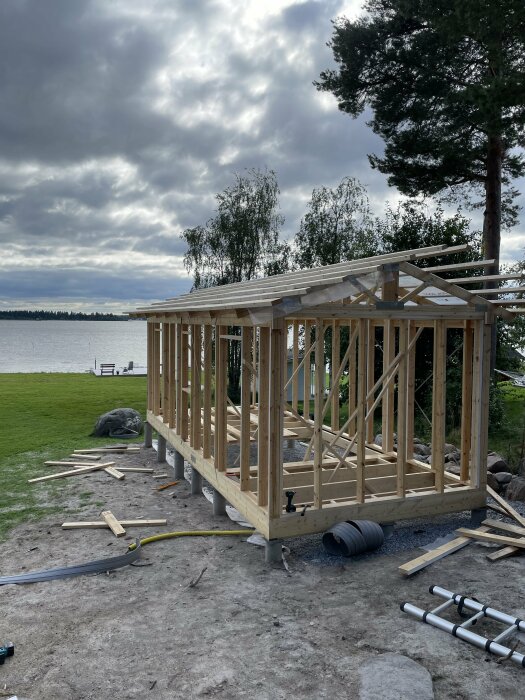 Trästomme under uppbyggnad nära en sjö, med väggblock och takstolar. Material och verktyg syns på marken omkring bygget.