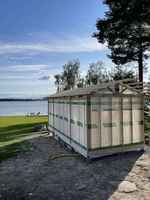 Byggstomme med väggblock och takstolar uppsatta nära en sjö, omgiven av grön natur och några träd under en klarblå himmel.