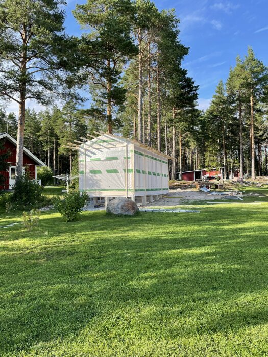 Nyuppställd byggnad med väggblock och takstolar, omgiven av grönska och tallar. Ligger på tomt med gräsmatta, med röda stugor i bakgrunden.